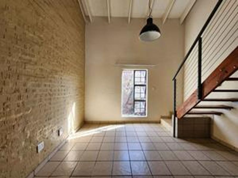 An empty living area featuring an exposed brick wall, tiled flooring, and a staircase with a metal railing under an exposed beam ceiling.