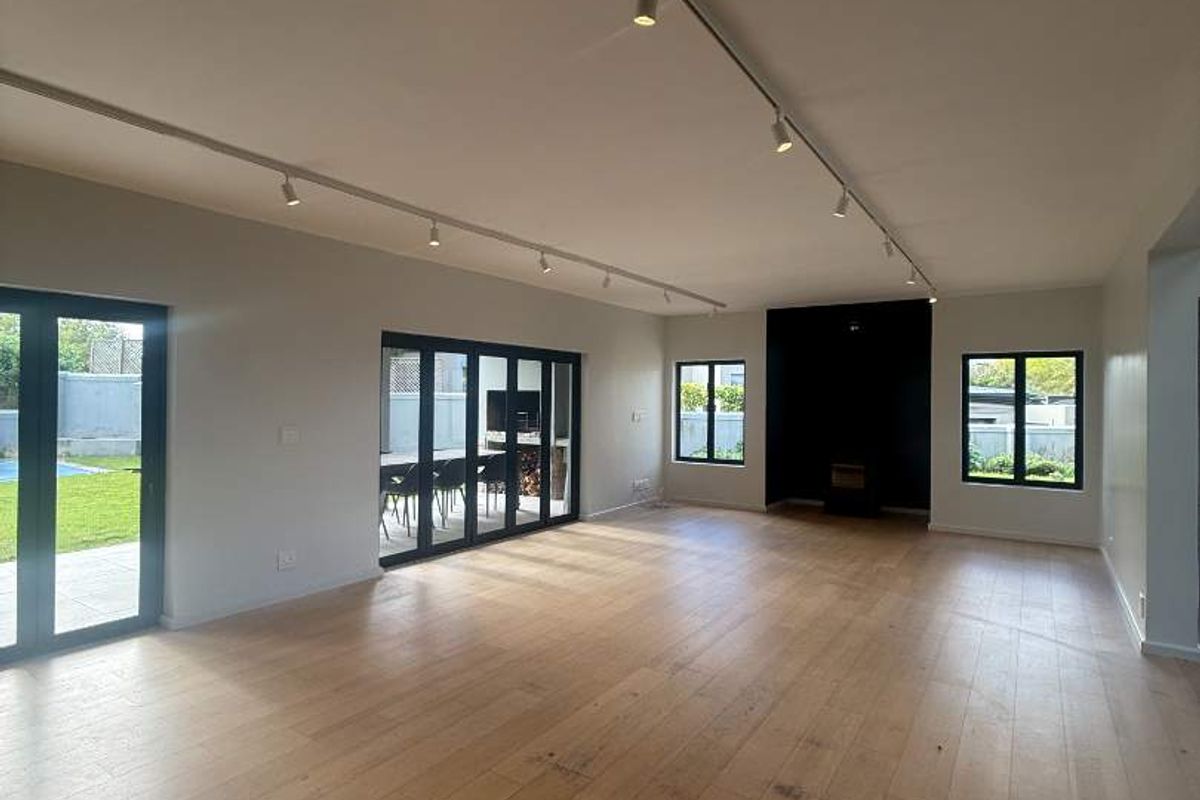 Spacious, light-filled living area featuring modern track lighting, light hardwood flooring, and large black-framed glass doors opening to the garden.