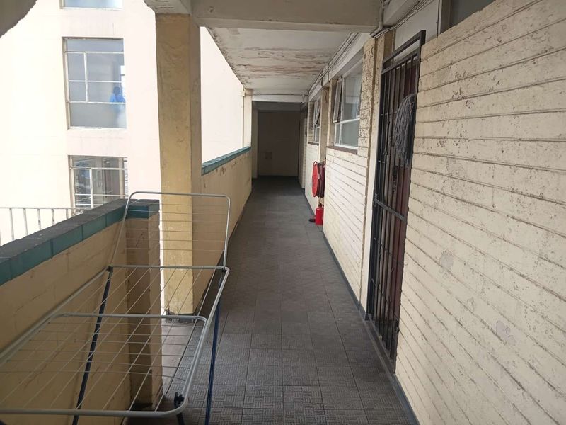 An exterior apartment building corridor featuring tiled flooring, a security gate, and a fire extinguisher, showing signs of wear on the ceiling and walls.