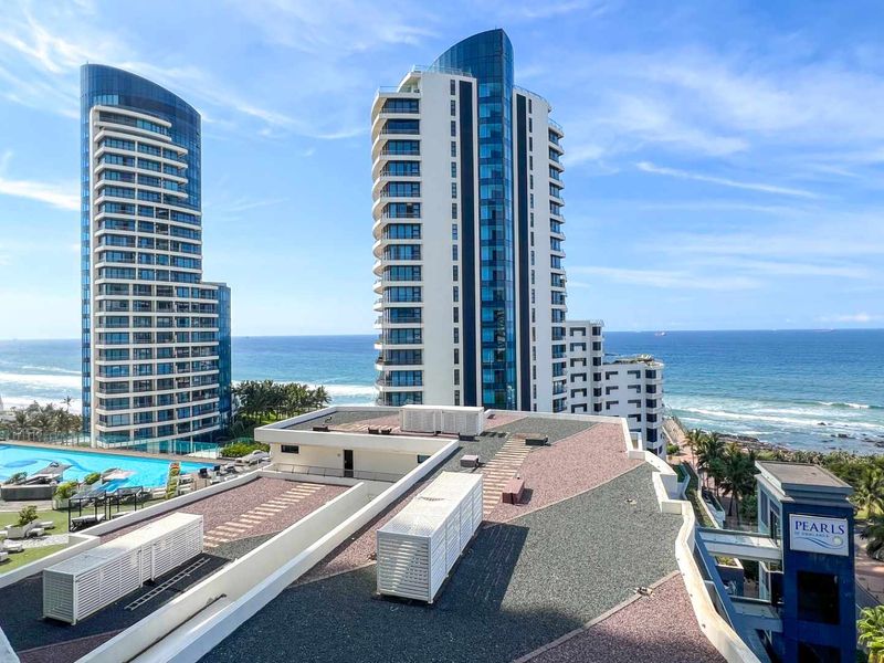 Elevated view overlooking modern high-rise apartment towers, a swimming pool area, and the coastline with ocean views.