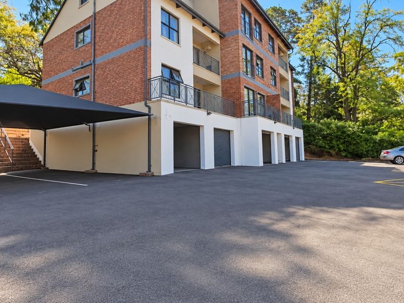 Exterior view of an apartment building featuring brick facade, private balconies with metal railings, ground-level garage access, and a paved parking area with shade cloth.