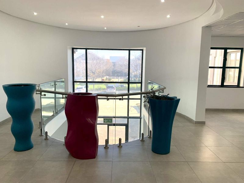A bright and spacious office hallway featuring modern tiled flooring, a glass balustrade overlooking the entrance, and large windows providing ample natural light.