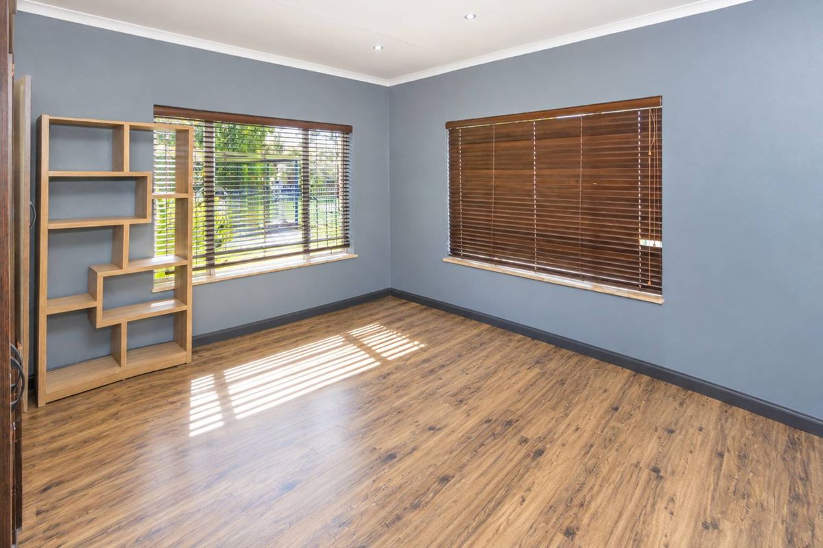 A bright, empty bedroom featuring modern grey walls, wood-look laminate flooring, and fitted wooden venetian blinds.