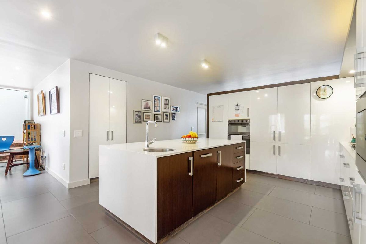 Modern kitchen featuring a central island with a sink, sleek white cabinetry, integrated oven, and large format floor tiles.