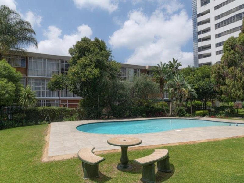 Communal outdoor pool area featuring a paved surround, well-maintained lawn, and established garden landscaping within an apartment complex setting.