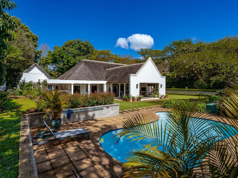 Rear view of a well-maintained house featuring a thatched roof, a covered patio area, and a swimming pool set within a landscaped garden.