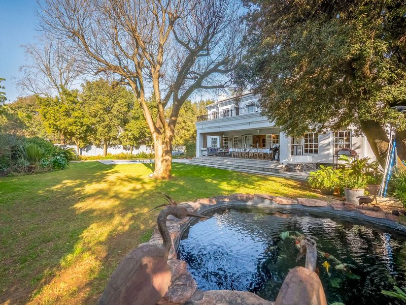 Spacious rear garden featuring a stone-lined pond, mature trees, and a covered patio leading to a two-story residence with a balcony.