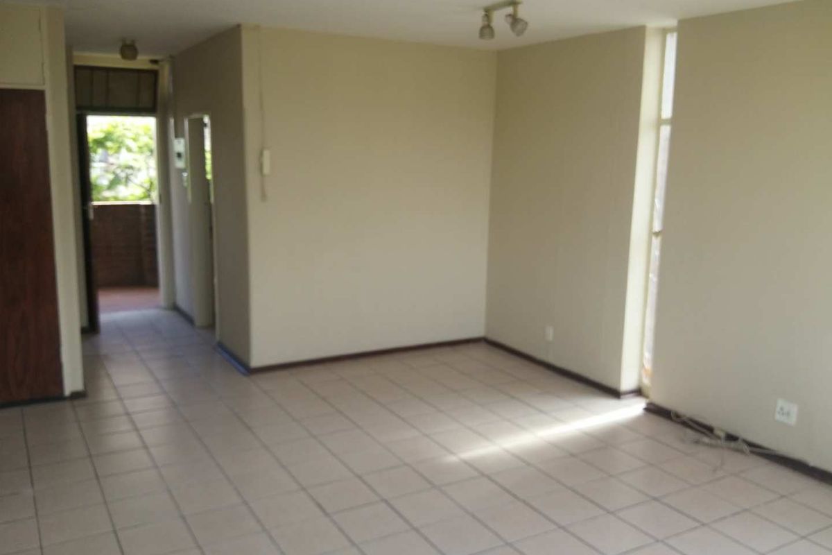 An empty, open-plan living area featuring neutral tiled flooring, ceiling spotlights, and a large window providing natural light.