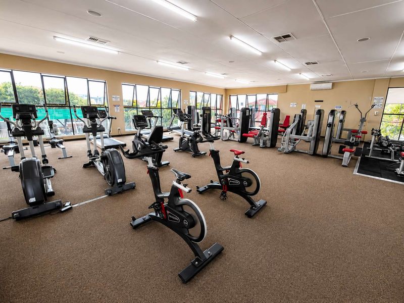 A spacious, well-lit communal gym facility featuring large windows, carpeted flooring, and a variety of fixed exercise equipment.
