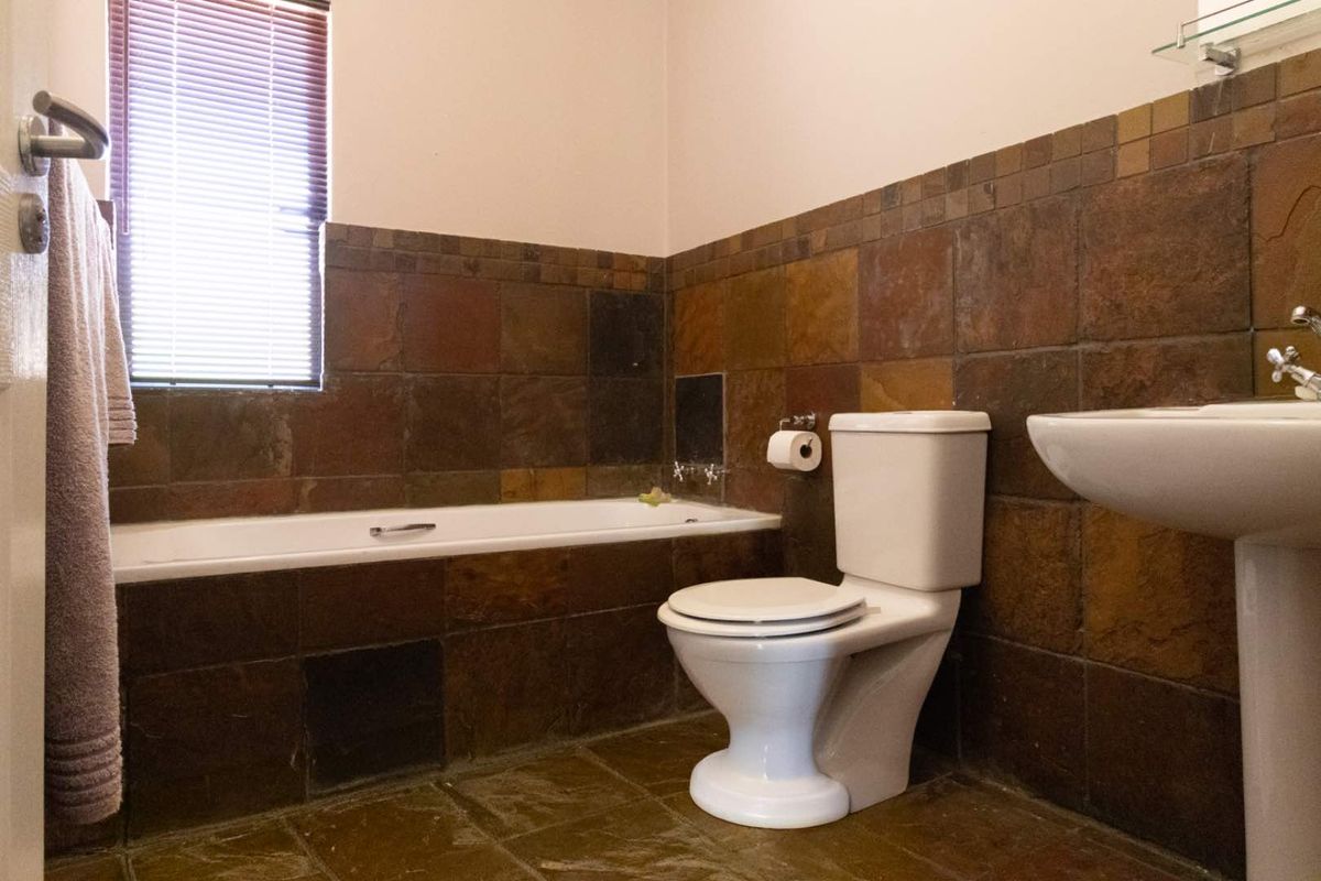 A functional bathroom featuring a built-in bathtub, pedestal basin, and toilet, finished with dark natural stone-style wall and floor tiling.