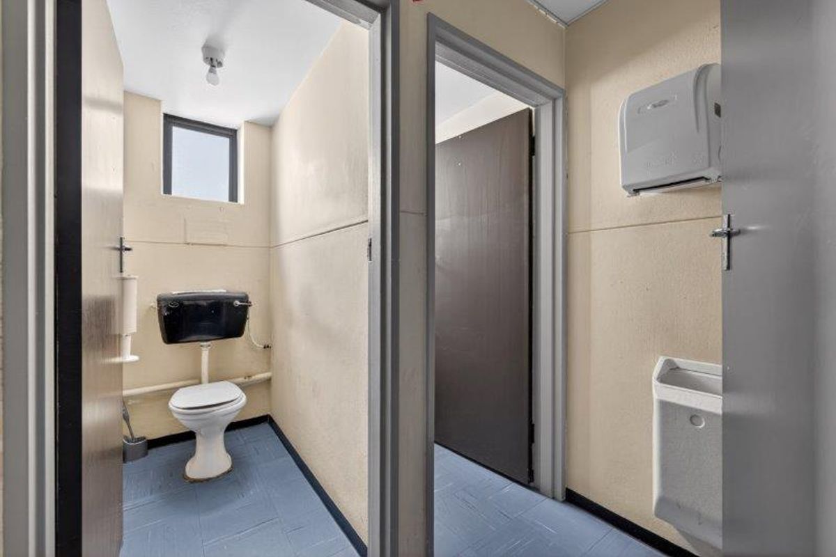 A functional office restroom facility featuring a toilet, wall-mounted urinal, and paper towel dispenser with durable vinyl flooring.