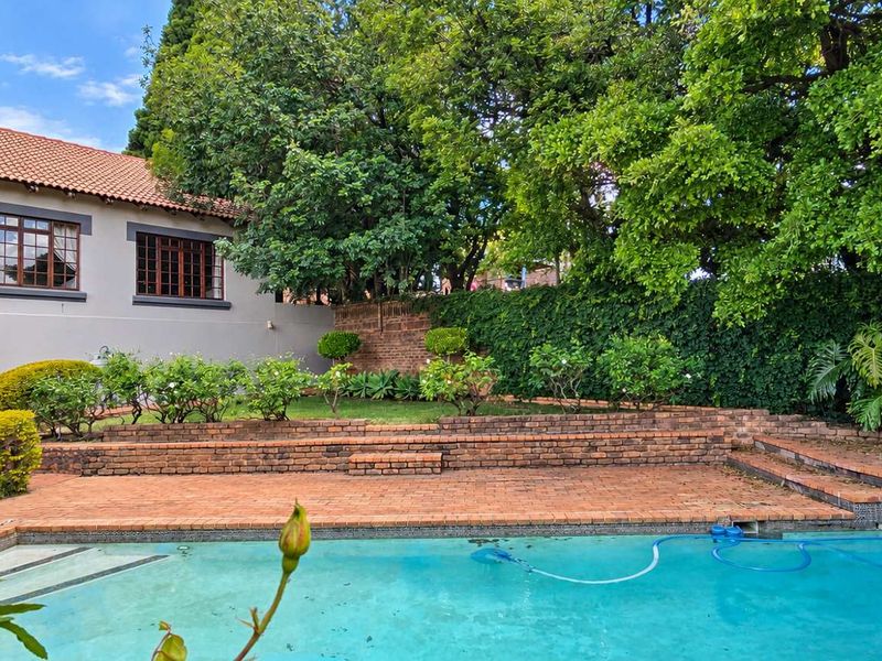 A private backyard featuring a swimming pool, multi-level brick paved patio, and established garden landscaping with mature trees.