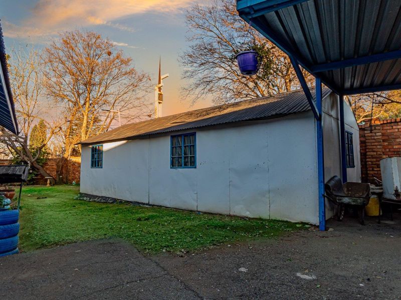 Rear exterior view showing a detached structure with corrugated metal roofing, a covered carport area, and a brick boundary wall in the garden.