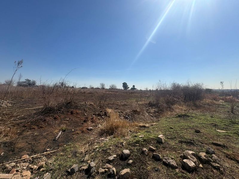 A wide-angle view of a vacant residential plot featuring natural, unlandscaped terrain with dry vegetation and scattered rocks under a clear sky.