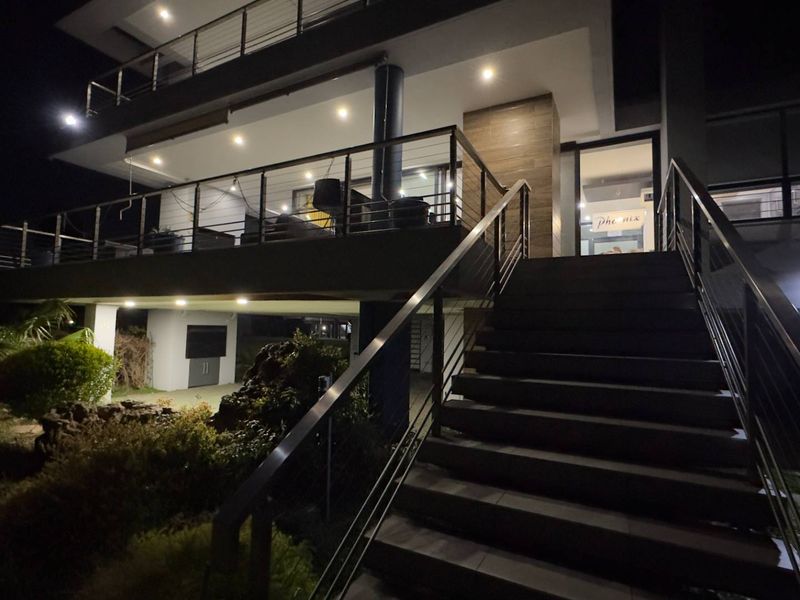 A modern multi-level home exterior at night, featuring a prominent staircase leading to an elevated patio with stainless steel railings and integrated lighting.