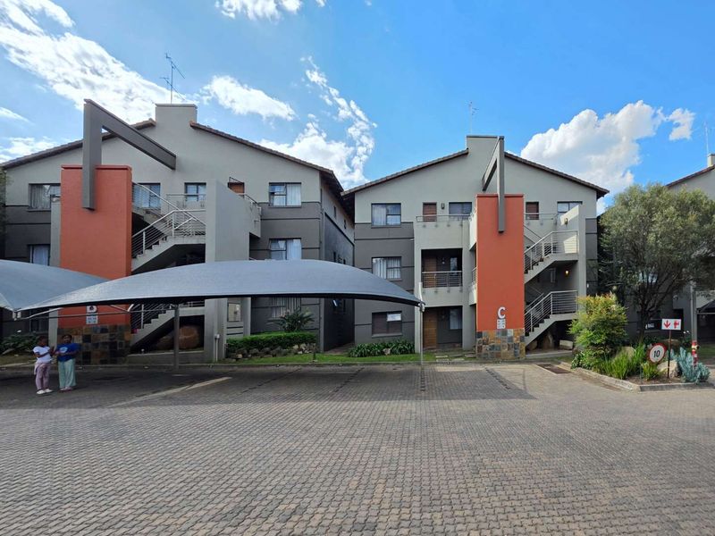 Front view of a multi-story apartment complex featuring paved parking, shade cloth carports, and modern architectural details with stone cladding accents.