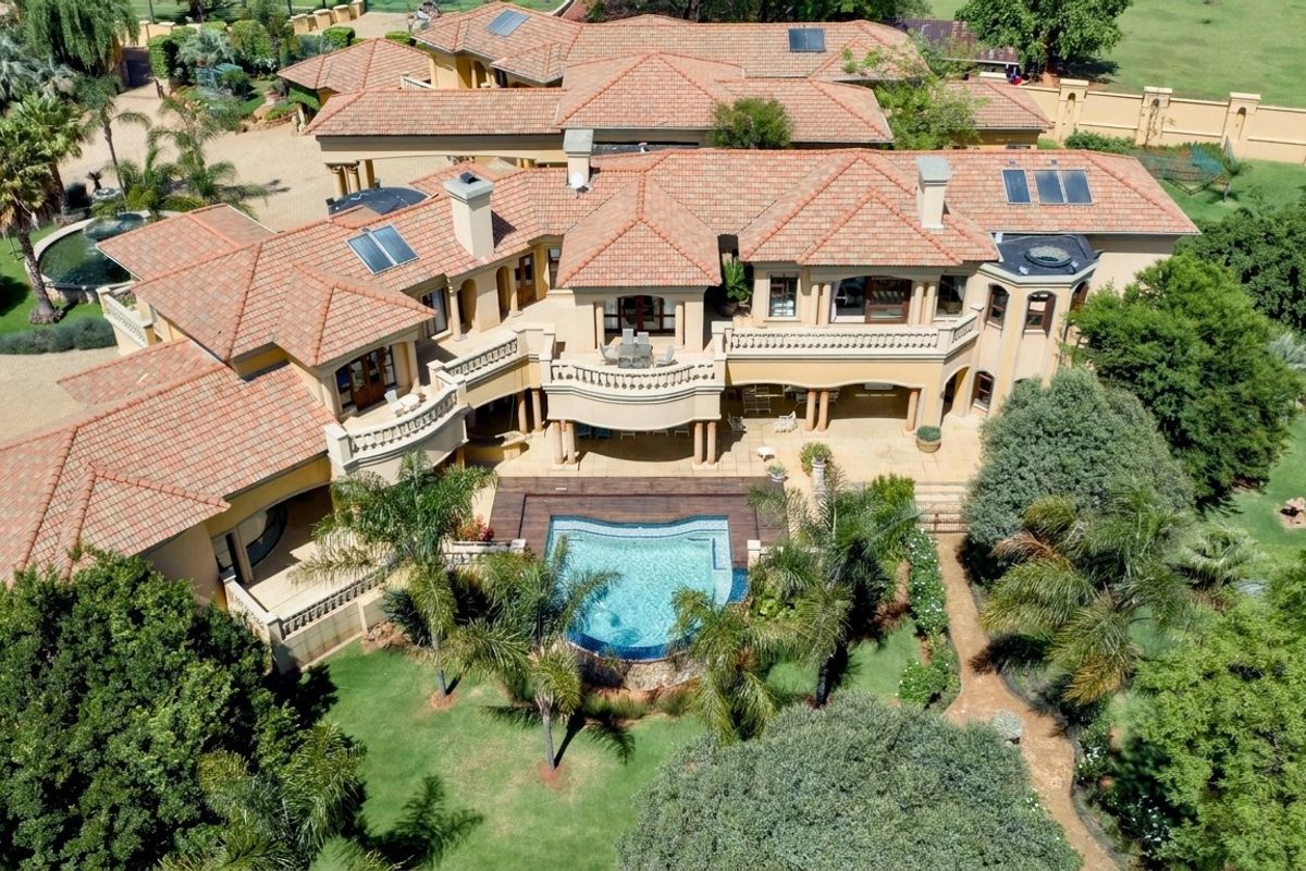 Aerial view of a large luxury residence featuring a terracotta tile roof, multiple balconies, a swimming pool with wooden decking, and extensive landscaped grounds.