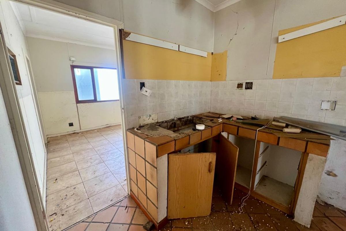 A dilapidated kitchen area requiring significant renovation, featuring original tiled counters, damaged wall finishes, and an adjacent room with tiled flooring.