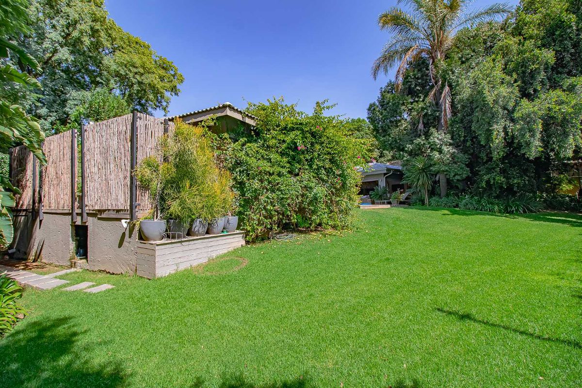 A spacious, well-maintained rear garden featuring a lush green lawn, mature trees, and a raised wooden deck area with privacy screening.
