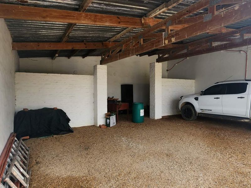 A spacious, rustic garage featuring exposed timber roof trusses, painted brick walls, and a gravel floor, providing ample covered parking or storage space.