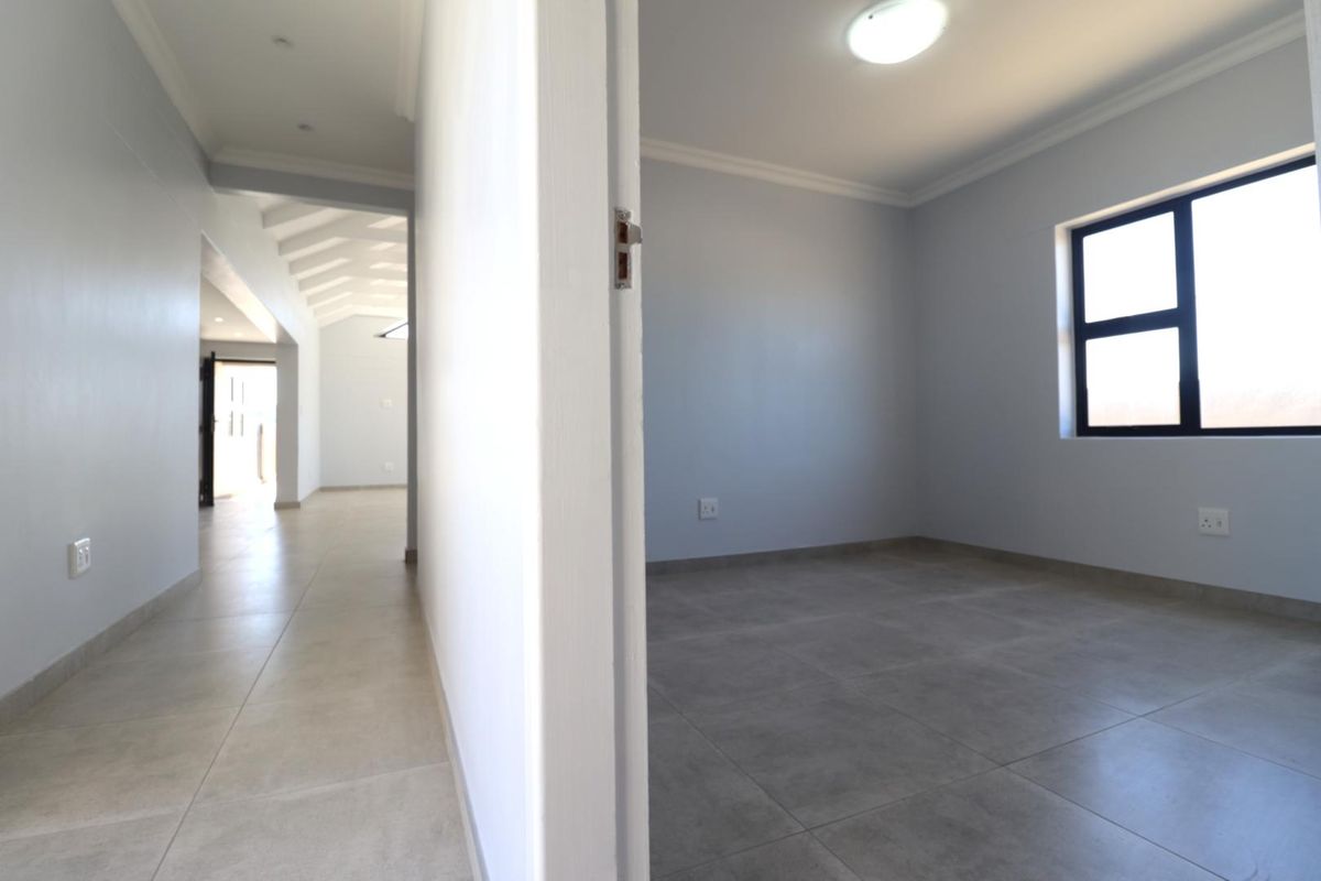 A bright, empty bedroom featuring neutral tiled flooring, modern window frames, and clean finishes, leading into a spacious hallway.