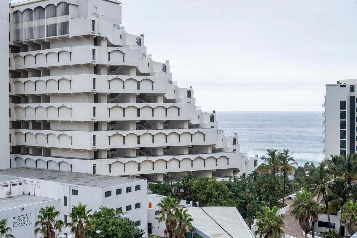 A view of a stepped apartment building facade featuring arched balconies, overlooking coastal grounds and the ocean.