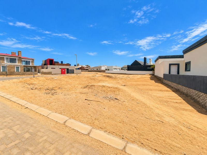 A vacant residential plot featuring a level sandy surface, paved curb, and established boundary walls in a developed neighborhood.