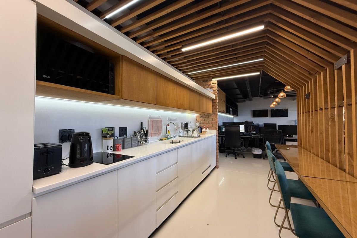 Modern office kitchenette featuring sleek white cabinetry, integrated appliances, and a unique timber-slatted ceiling design, seamlessly connecting to an open-plan workspace.