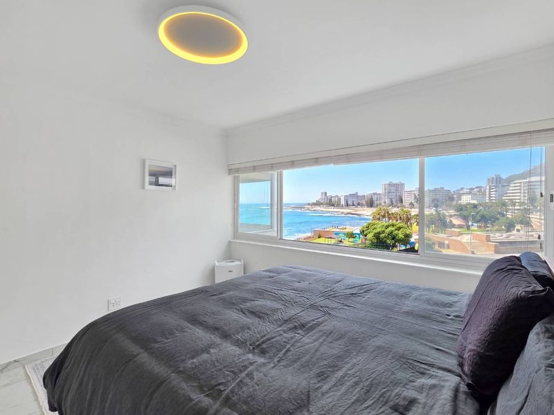 A bright bedroom featuring a large picture window with expansive coastal and city views, complemented by modern lighting and clean finishes.