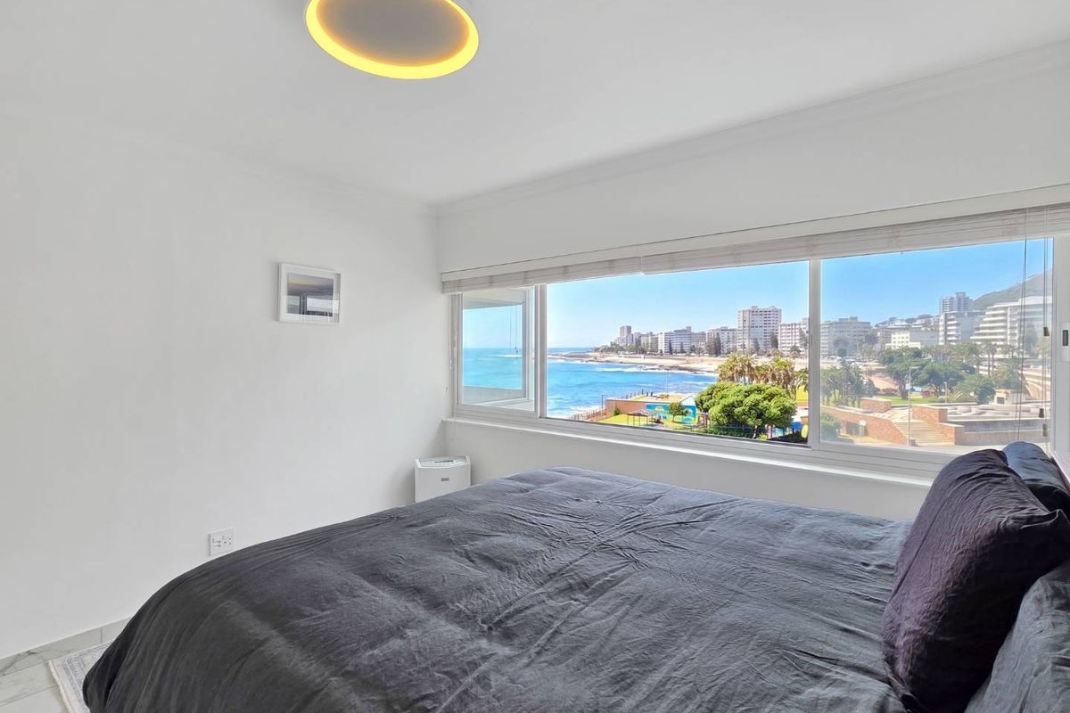 A bright bedroom featuring a large picture window with expansive coastal and city views, complemented by modern lighting and clean finishes.