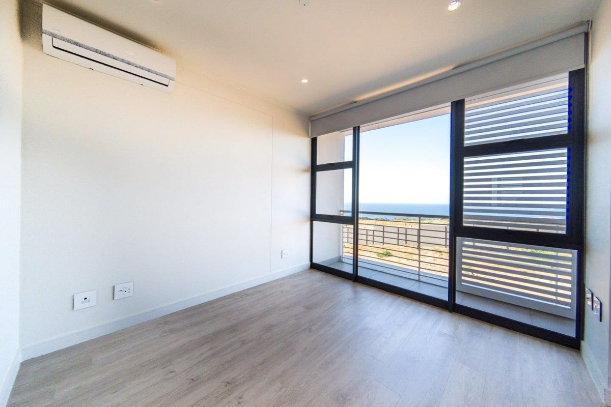 A bright, empty bedroom featuring modern laminate flooring, a split-system air conditioner, and large sliding glass doors leading to a balcony with ocean views.