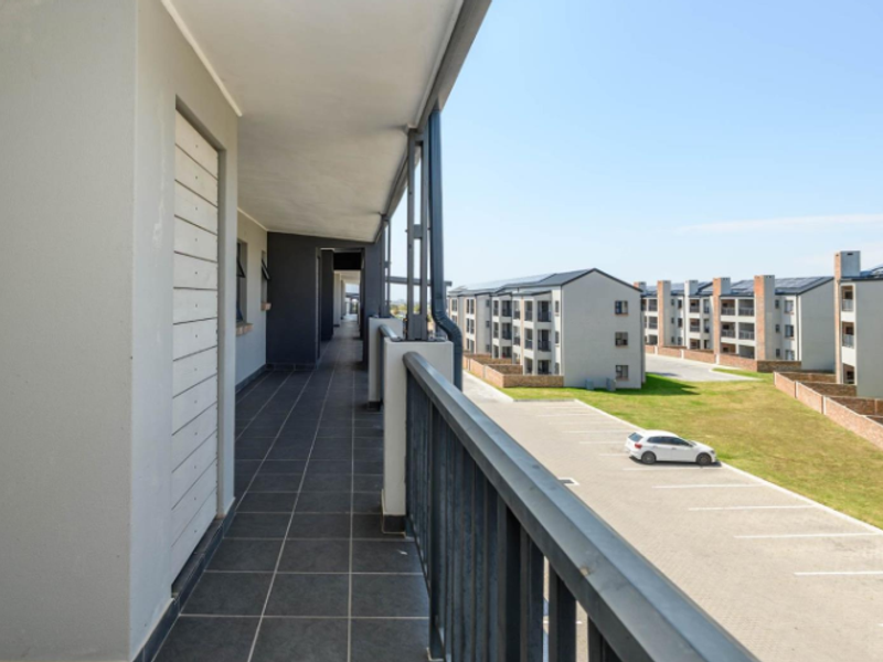 A spacious covered balcony with tiled flooring and a metal balustrade, overlooking the apartment complex grounds and parking area.