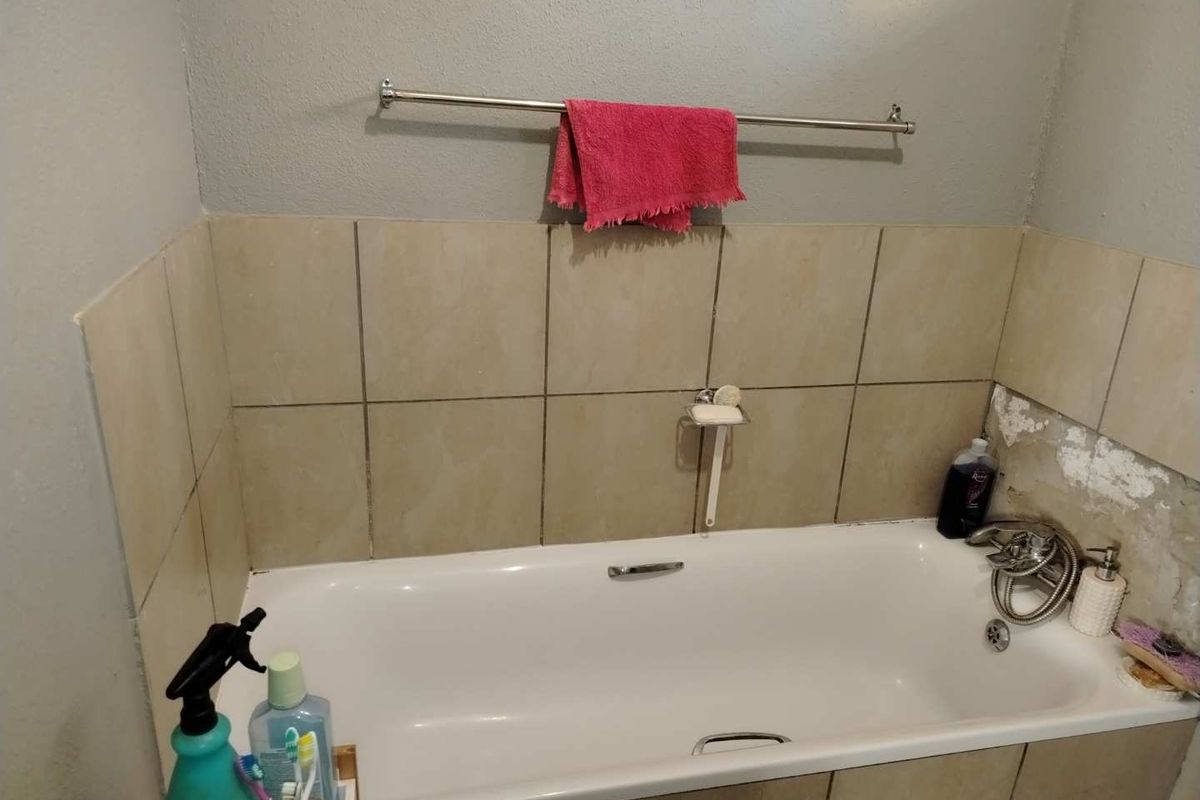 A bathroom featuring a standard bathtub with tiled surround, chrome fixtures, and a towel rail, currently showing signs of wall surface damage.