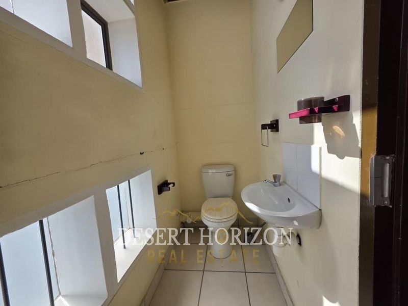 A compact industrial-style bathroom featuring a pedestal washbasin, toilet, and tiled flooring, requiring minor cosmetic repairs to the wall surfaces.