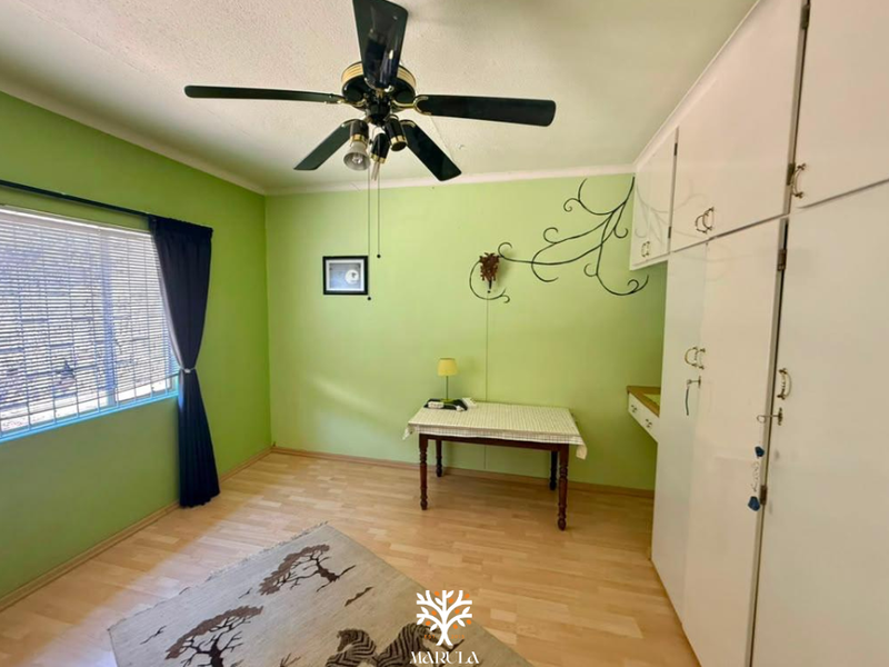 A bedroom featuring laminate flooring, built-in cupboards, and a ceiling fan, with large windows fitted with venetian blinds.