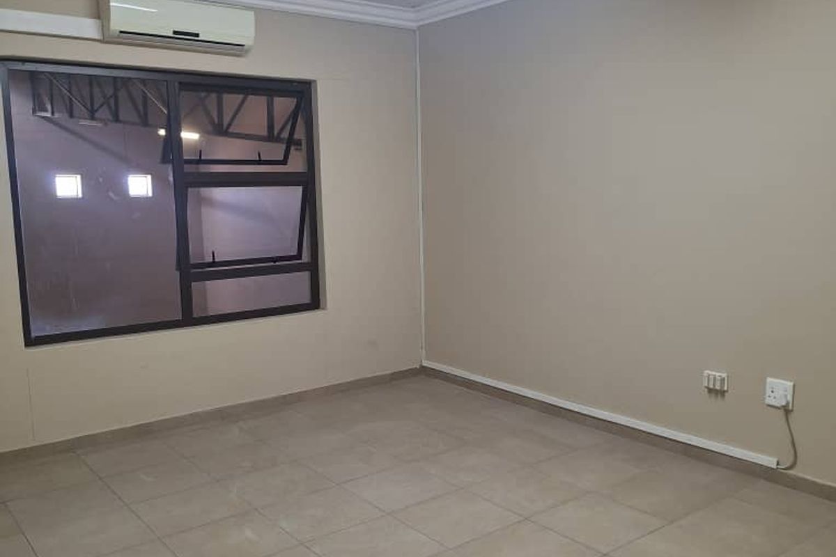 A clean, empty room featuring neutral tiled flooring, a wall-mounted air conditioning unit, and a large window with security bars.