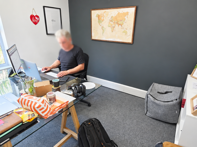 A functional office space featuring grey carpet flooring, a dark accent wall, and natural light provided by a large window with venetian blinds.