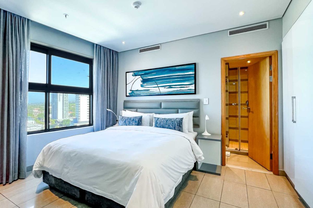 A modern bedroom featuring tiled flooring, built-in wardrobes, large windows with city views, and an en-suite bathroom with a glass-enclosed shower.