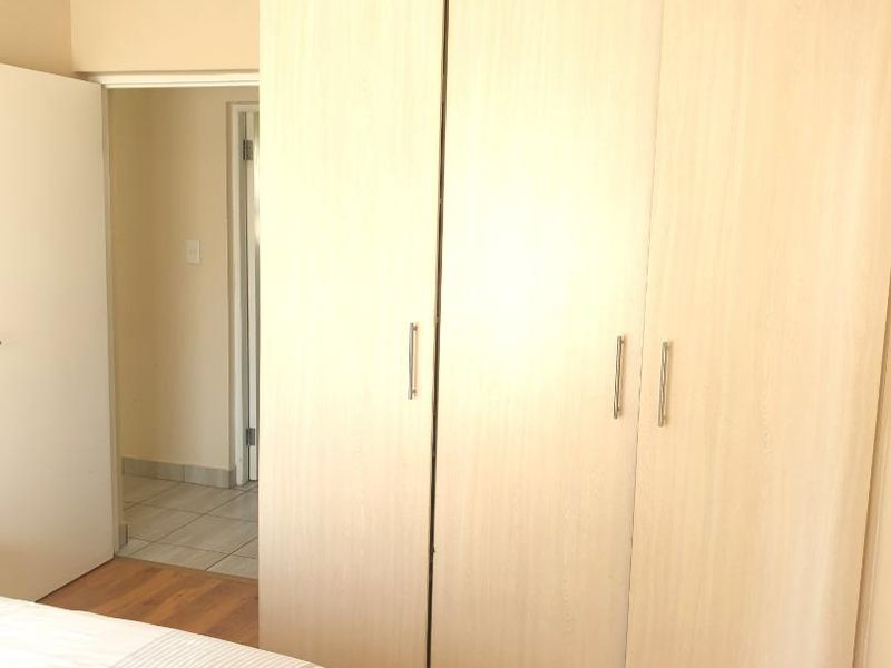 A bedroom featuring light wood-grain built-in cupboards and warm-toned laminate flooring, with an adjacent doorway leading to a tiled hallway.