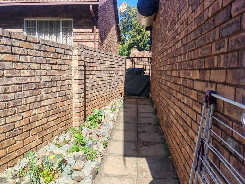 A narrow side passage featuring a brick boundary wall, paved walkway, and a small rock garden bed leading to a rear security gate.