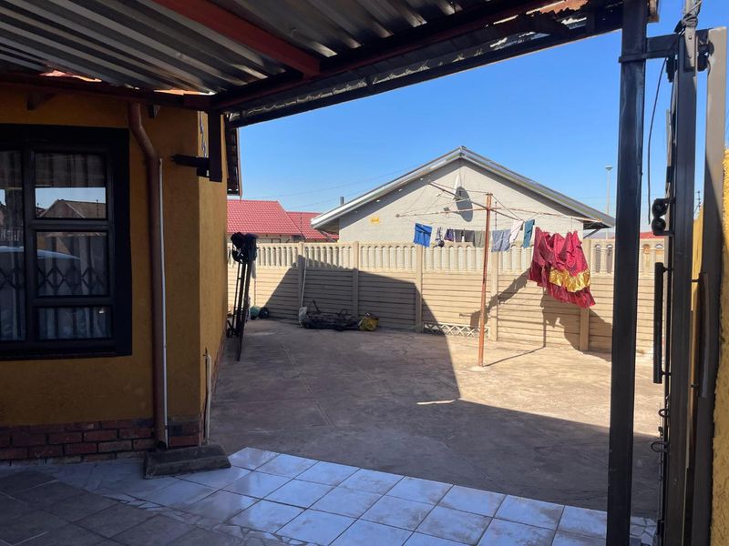 A covered outdoor patio area featuring tiled flooring and a corrugated metal roof, overlooking a paved backyard with a precast concrete boundary wall.
