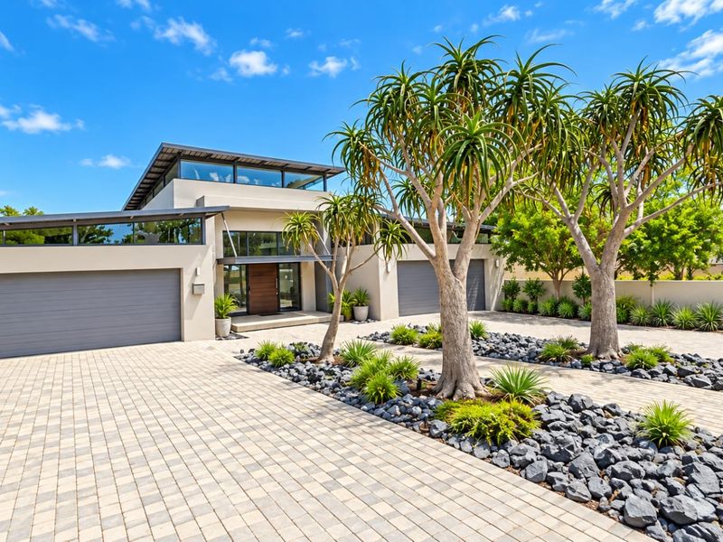 A modern architectural home featuring a paved driveway, double garage, and well-maintained landscaped garden with mature trees and stone accents.