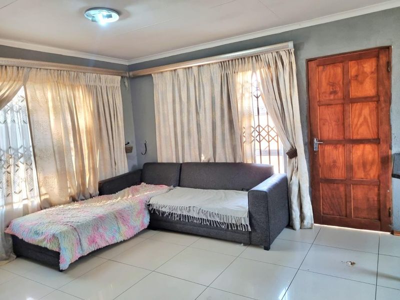 A living room featuring tiled flooring, a wooden entrance door with security gate, and large windows with curtain tracks.