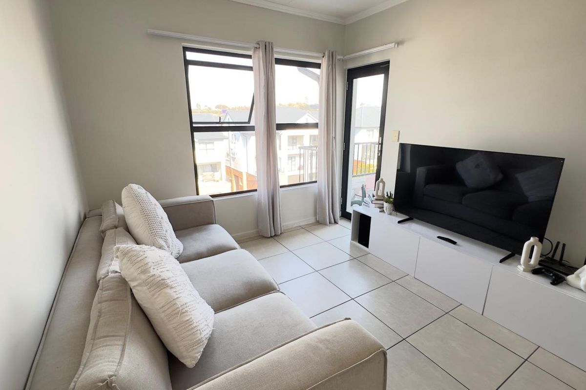 A bright living room featuring neutral tiled flooring, large aluminium-framed windows, and direct access to an outdoor balcony area.