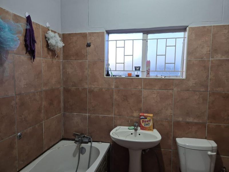 A functional bathroom featuring a pedestal basin, bathtub, and toilet, finished with brown wall tiling and security bars on the window.