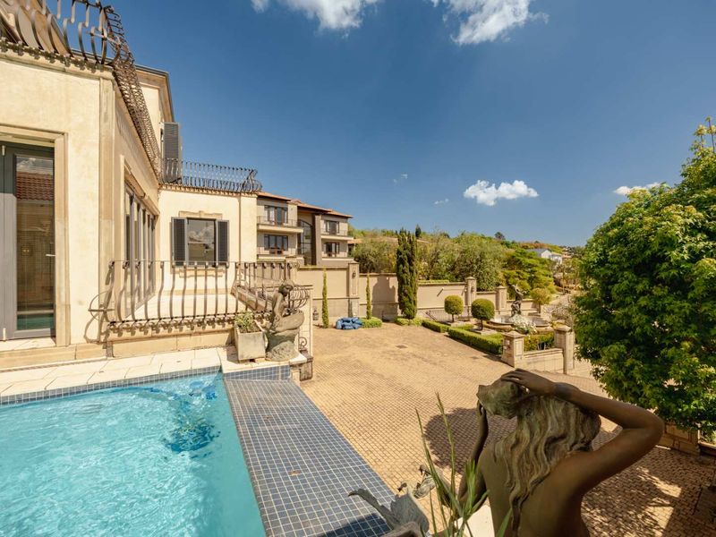 A beautifully maintained outdoor pool area featuring a tiled swimming pool, wrought iron railings, and a spacious paved courtyard with landscaped garden elements.