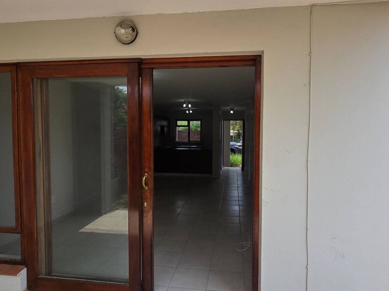 Covered patio area featuring a wooden sliding door entrance leading into the main living space, with tiled flooring and exterior wall lighting.