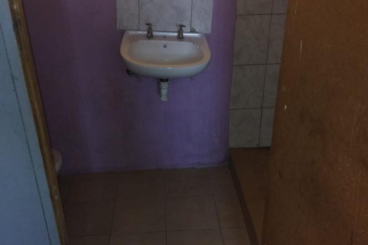 A compact bathroom featuring a wall-mounted basin, tiled flooring, and security bars on the window, requiring cosmetic updates to modernize the space.
