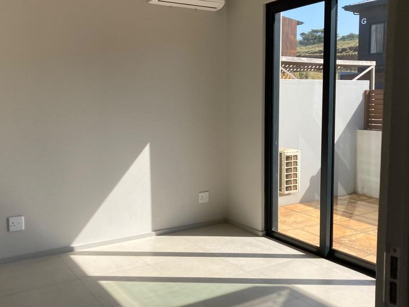 A bright, empty living room featuring neutral tiled flooring, a split-system air conditioner, and large sliding glass doors leading to an outdoor patio area.