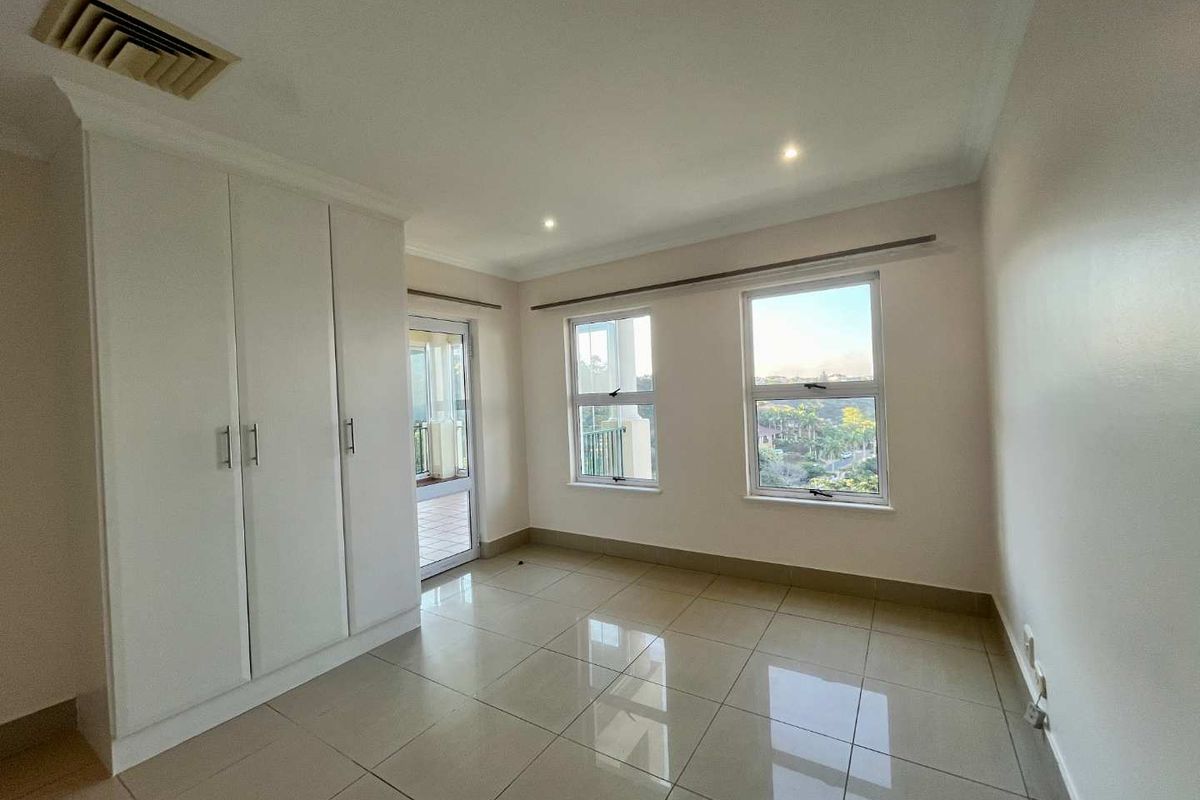 A bright, empty bedroom featuring tiled floors, built-in cupboards, recessed lighting, and direct access to a private balcony through a glass door.